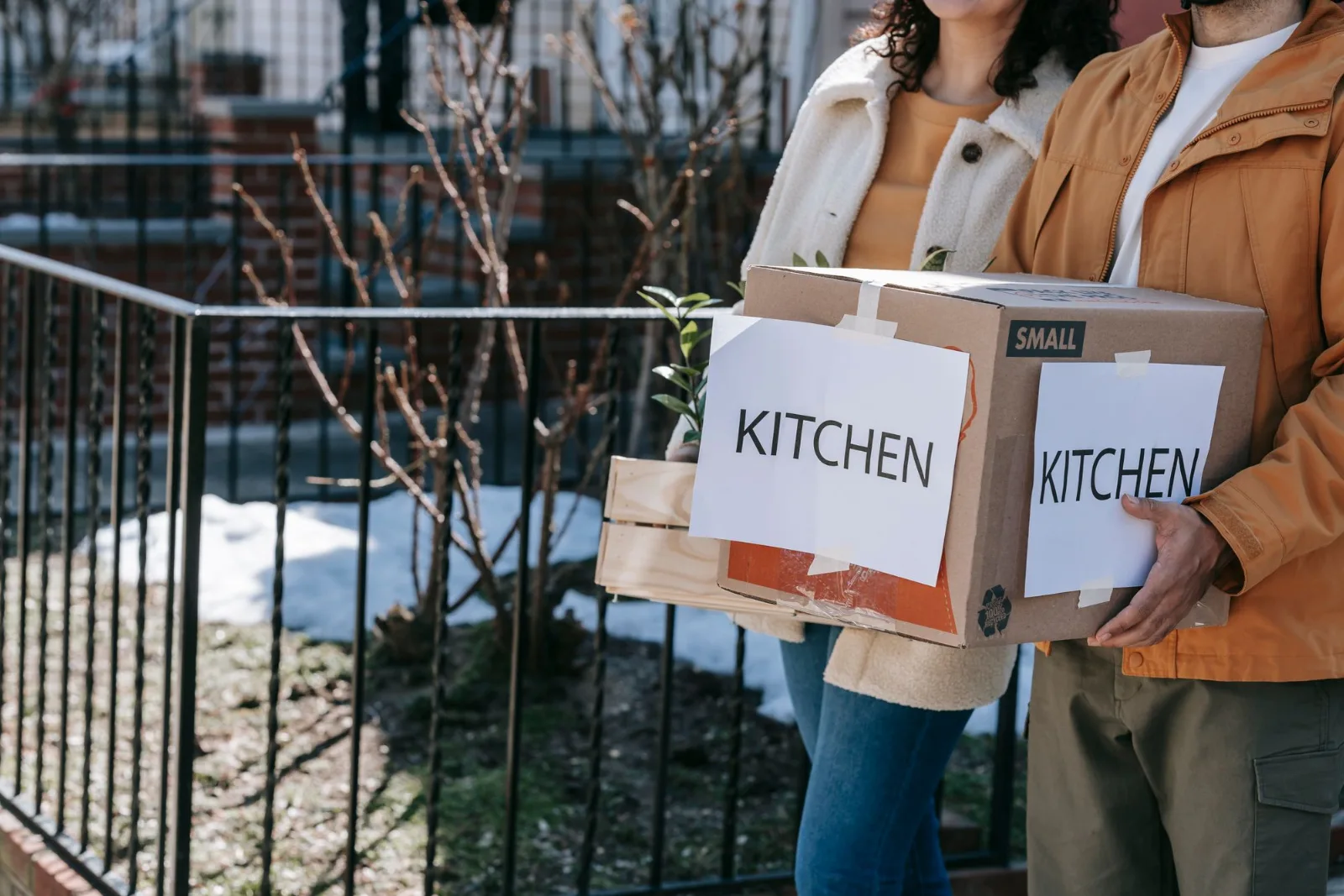 Two people carrying labelled kitchen boxes outdoors while moving into a new home