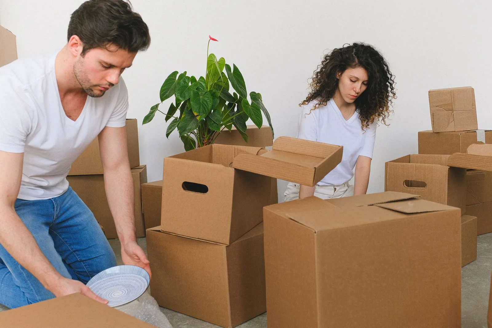 Two people wrapping fragile items and packing them into a cardboard carton in a bright room