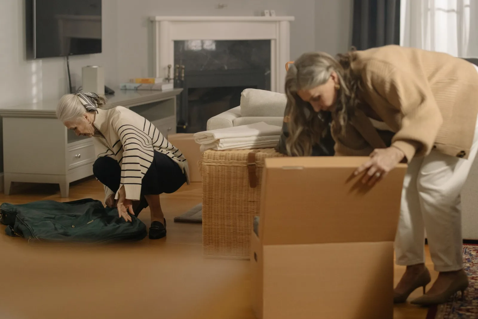 Two older women packing belongings into boxes in a living room