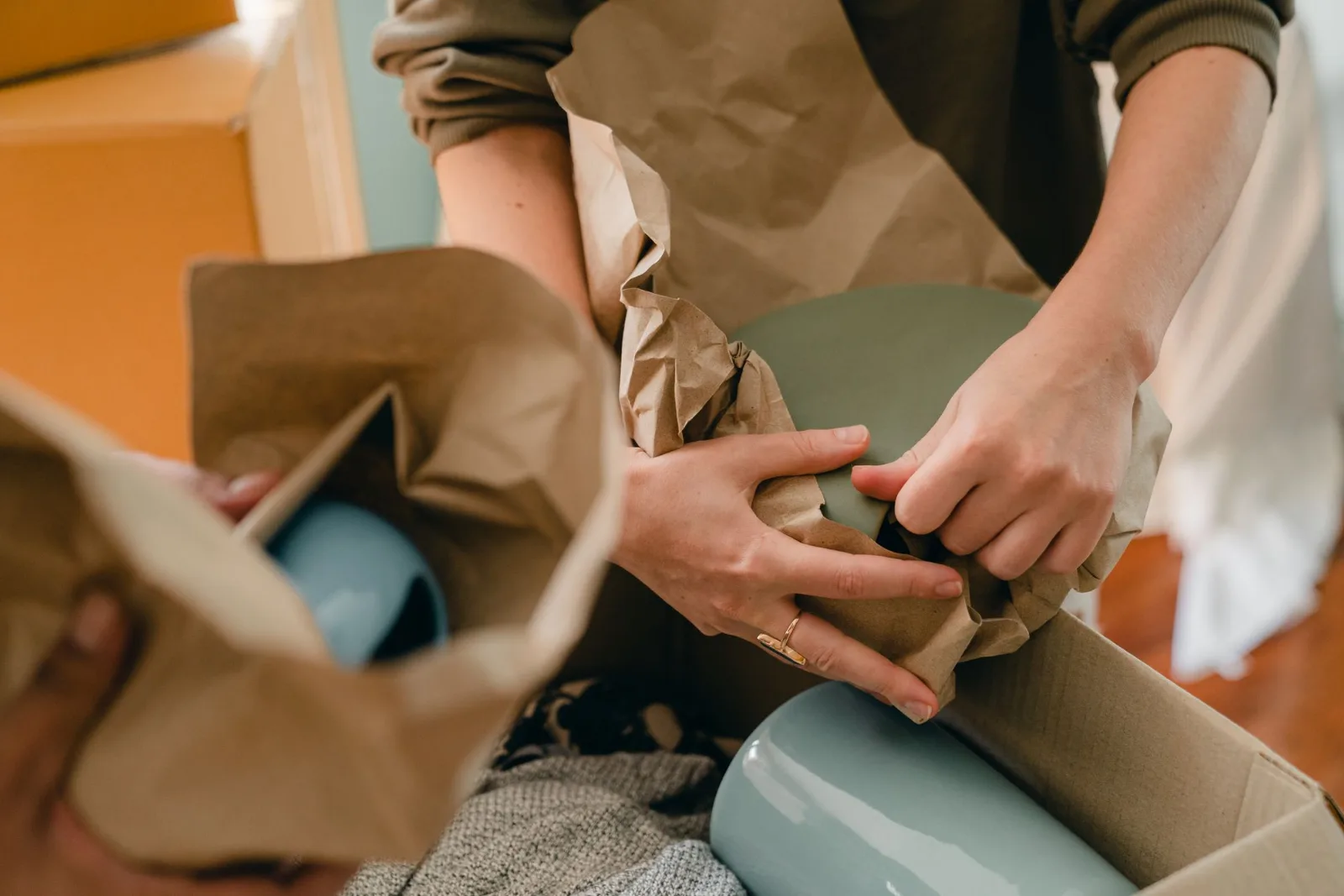 Hands wrapping a ceramic plate in clean packing paper beside a stack of dishes